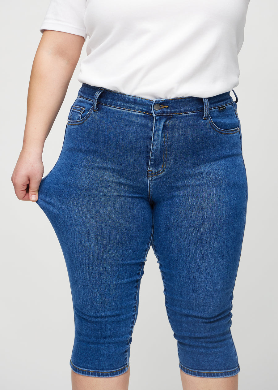 Skinny capris for women in dark blue denim, color Oceans. A plus-size model stretches the fabric to demonstrate its flexibility.