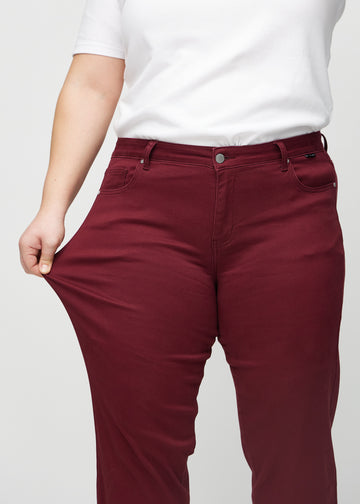 Regular jeans for women in dark red, color Santas. A plus-size model stretches the fabric at the thigh to show its stretch.