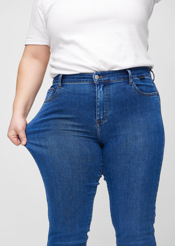 Slim jeans for women in dark blue, color Oceans. A plus-size model stretches the jeans at the thigh to show the fabric&