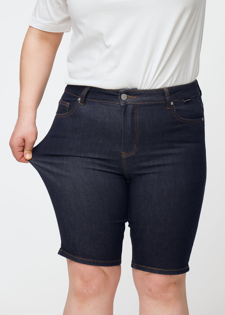 Plus-size model stretches deep dark blue denim shorts for women in the colour Midnights at the thigh to show stretch.