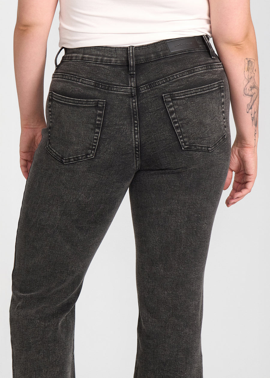 Mørkegrå denim bootcut jeans, modelnavn Shadows, som sidder tæt om hofter og lår med et let svaj fra knæet og ned, set bagfra tæt på for at vise detaljer. på en plus-size model.